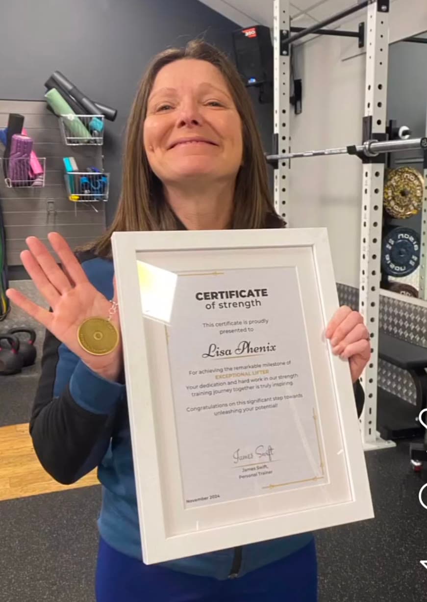 Lisa smiling, holding her framed Certificate of Strength and custom medal for reaching intermediate strength milestones